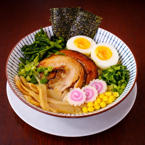 A vibrant bowl of ramen with chashu pork, soft-boiled eggs, nori, narutomaki, and fresh greens.