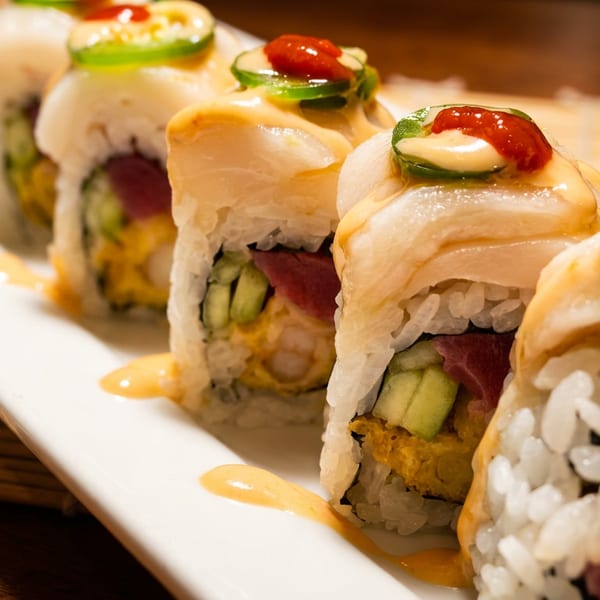 Sushi rolls topped with white fish, creamy sauce, jalapeño, and red chili sauce on a white plate.